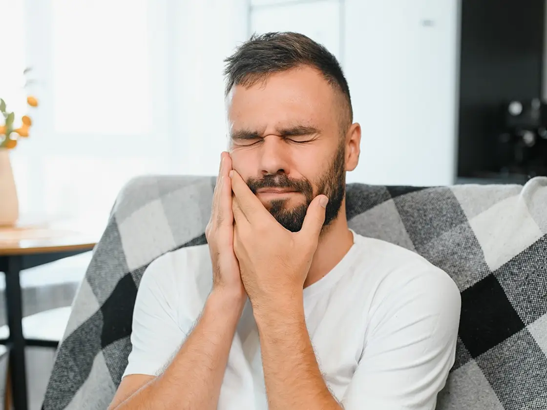 A man sitting on a sofa at home holding his jaw in discomfort