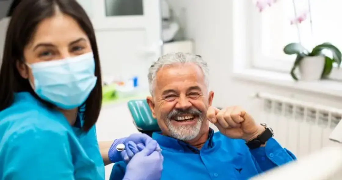 A smiling older patient giving a thumbs up in the dental chair beside a masked dental assistant