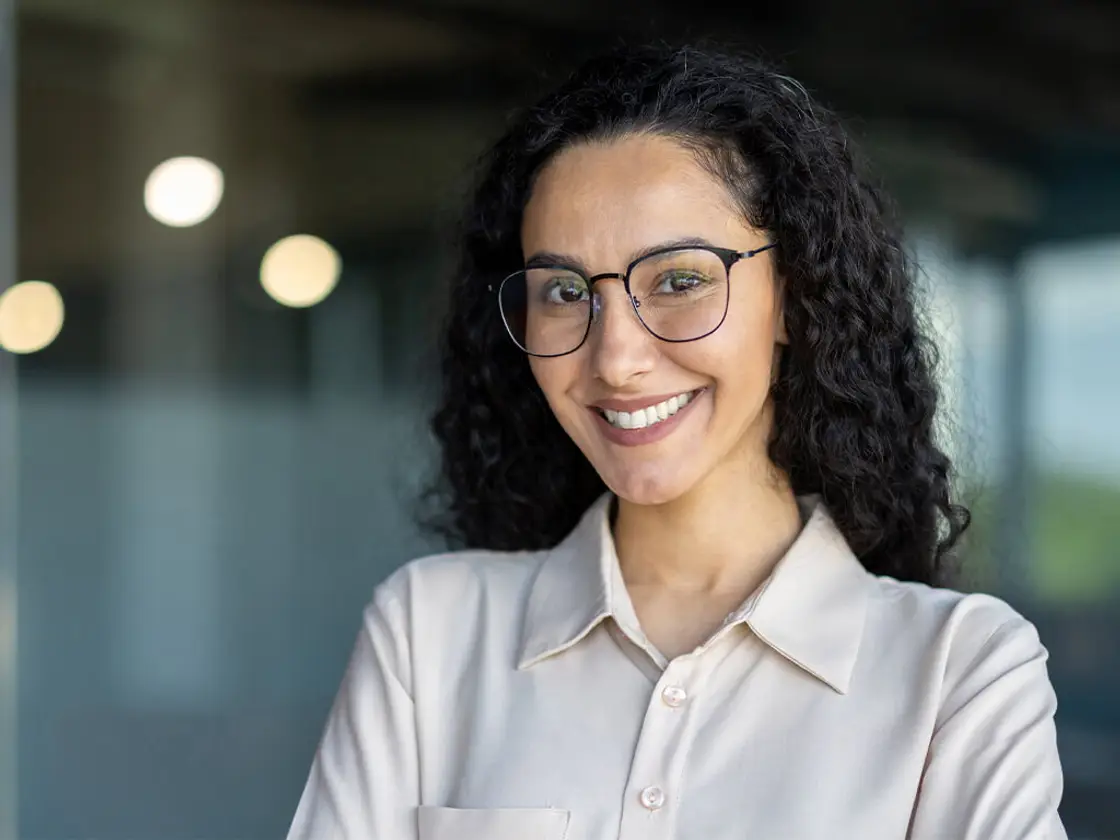 A woman in glasses with a bright, even smile standing with her arms folded