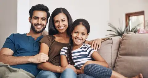 A family of three sitting together on a living room sofa, smiling at the camera